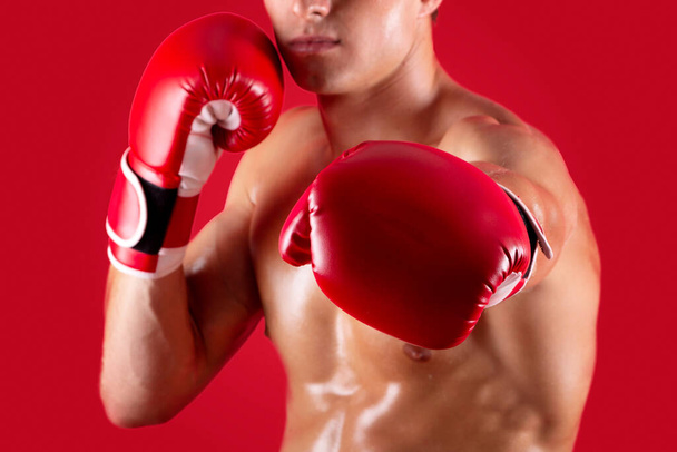 Determined young boxer in fighting gloves making punch at camera on red studio background, selective focus - Valokuva, kuva