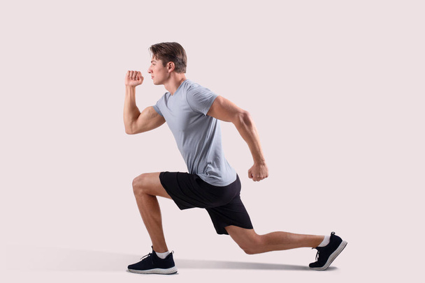 Side view of strong millennial guy in sportswear doing lunges on light studio background - Foto, Imagen