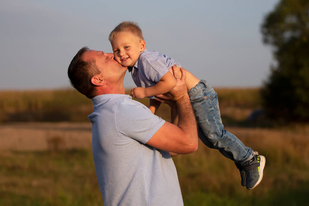 幸せなお父さんは畑の背景で彼の息子と遊ぶ。喜びの祖父は彼の孫にキス. - 写真・画像