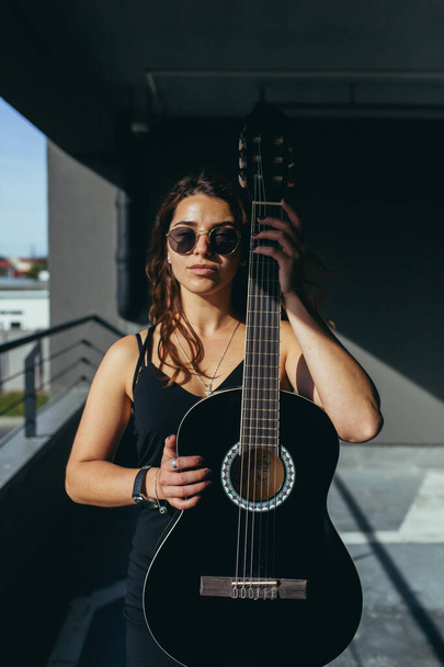 Beautiful rock and roll girl in black glasses posing with a black guitar in the car park - Foto, imagen