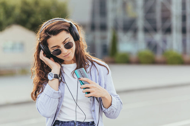Young pretty student girl, waiting for a taxi car, and listening to music from the phone on big headphones - Фото, изображение
