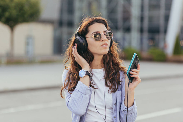 Young pretty student girl, waiting for a taxi car, and listening to music from the phone on big headphones - Fotoğraf, Görsel
