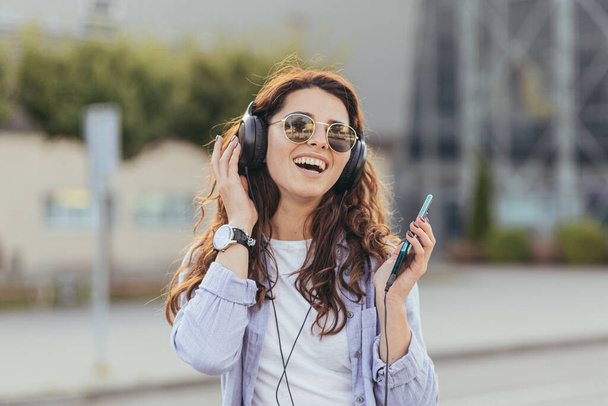 Young pretty student girl, waiting for a taxi car, and listening to music from the phone on big headphones - Фото, изображение