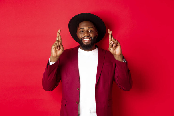 Hopeful african american man making wish, holding fingers crossed for good luck and smiling optimistic, standing against red party background - Foto, afbeelding