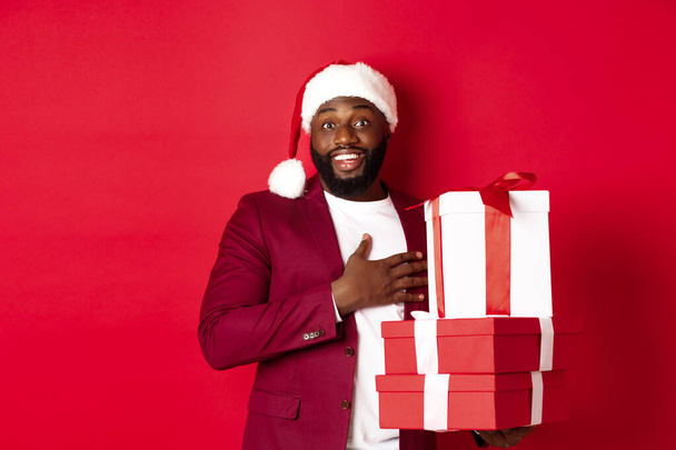 Joulu, uusi vuosi ja ostokset käsite. Happy Black mies vastaanottaa joululahjoja, kiittäen ja hymyillen kiitollinen, seisoo jouluhattu punaista taustaa vasten - Valokuva, kuva