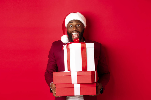 Noel, yeni yıl ve alışveriş konsepti. Noel Baba şapkalı ve ceketli mutlu siyah adam elinde Noel hediyeler, hediyeler ve gülücükler getiriyor, kırmızı arka planda duruyor. - Fotoğraf, Görsel