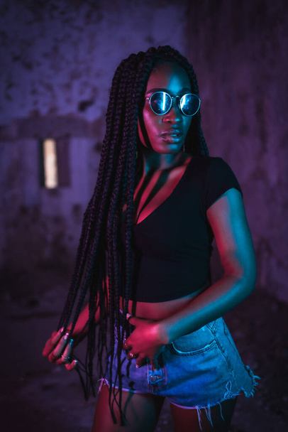 Lifestyle, portrait of a young black woman with long braids, sunglasses and a black t-shirt. Pink and blue neon lights, urban photography of a trap dancer in serious pose - Photo, Image