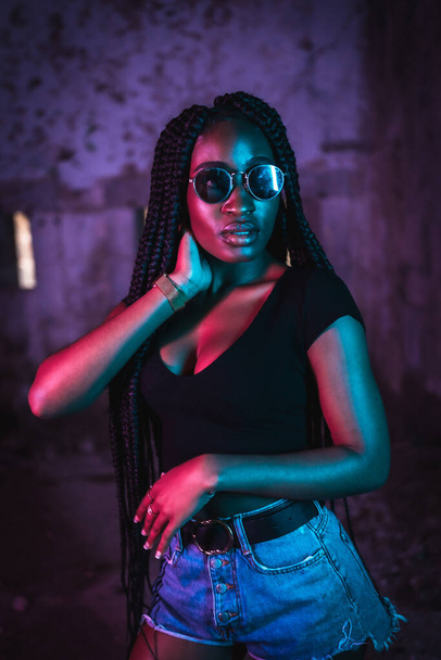 Lifestyle, portrait of a young black woman with long braids, sunglasses and a black t-shirt. Pink and blue neon lights, urban photography of a trap dancer - Photo, Image