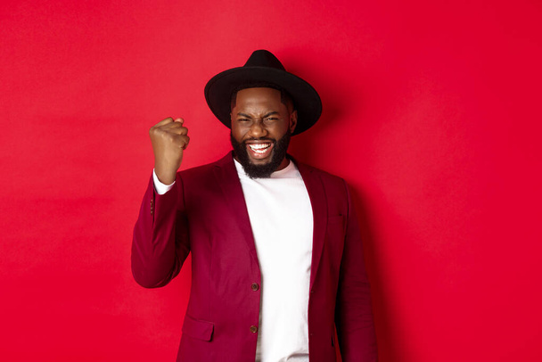 Fashion and party concept. Excited Black man rejoicing of winning, making fist pump gesture and say yes with satisfaction, standing against red background - Photo, Image