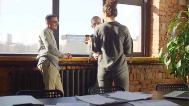 Mediana toma de joven mujer amigable trayendo café a sus compañeros de trabajo masculinos de pie junto a una gran ventana hablando y sonriendo descansando del trabajo a la luz del día - Metraje, vídeo