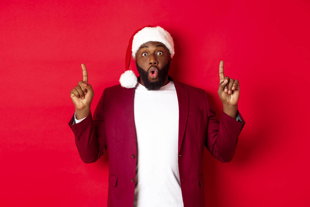Christmas, party and holidays concept. Impressed Black man showing offer, pointing fingers up at logo, saying wow, standing against red background - Fotó, kép