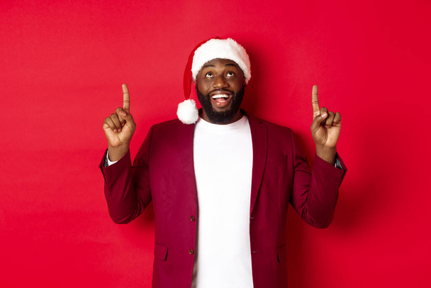 Concepto de Navidad, fiesta y vacaciones. Feliz hombre negro mirando la promoción de Año Nuevo, señalando con los dedos hacia arriba y sonriendo, de pie sobre el fondo rojo - Foto, Imagen