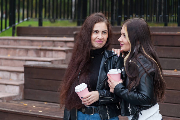 Kaksi parasta ystävää, nuori brunette naiset pitkät hiukset, seistä puistossa syksyllä taustaa vasten puinen penkki, pidä kuppeja kahvia käsissään ja toinen heistä halata toinen olkapää - Valokuva, kuva