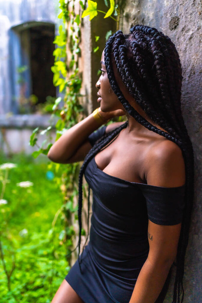 Lifestyle, Latina girl with black skin in a tight black dress and black heels. In an abandoned place full of nature looking at the landscape - Φωτογραφία, εικόνα