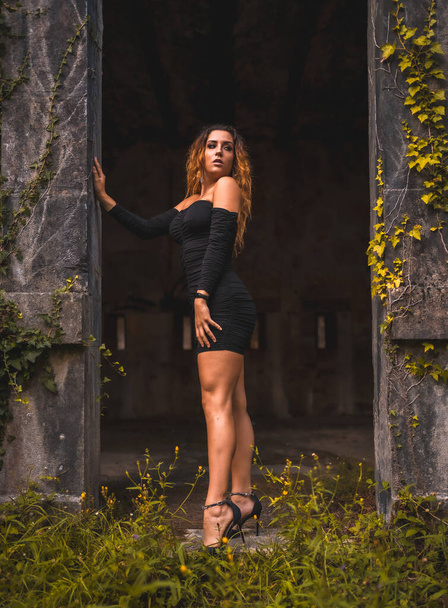 Lifestyle, Caucasian girl with wavy hair in a tight black dress and black high heels. Relaxed an abandoned place full of nature looking to the right - Foto, Imagen
