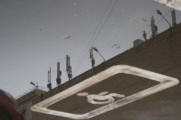 A handicap parking sign painted on the asphalt and the reflection of antennas on a building in a puddle next to it. Unusual angle - Foto, immagini
