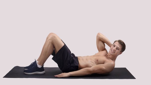 Smiling millennial guy with naked chest doing abs exercises on sports mat over light studio background, panorama - Photo, Image