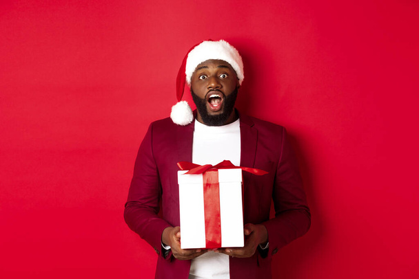 Natal, Ano Novo e conceito de compras. Homem negro animado recebendo presente de Natal, olhando surpreso, segurando presente embrulhado e olhando para a câmera, vestindo chapéu de Papai Noel - Foto, Imagem