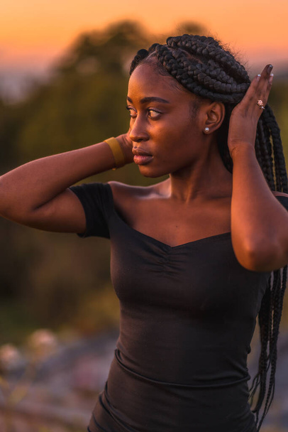 Lifestyle, Latina girl with black skin with beautiful long braids in her hair, wearing a tight black short dress. Portrait of young woman enjoying summer sunset in nature - Foto, Imagem