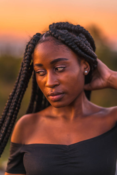 Lifestyle, Latina girl with black skin with beautiful long braids in her hair, wearing a tight black short dress. Portrait of the young man on top of the mountain in a beautiful summer sunset - Foto, afbeelding
