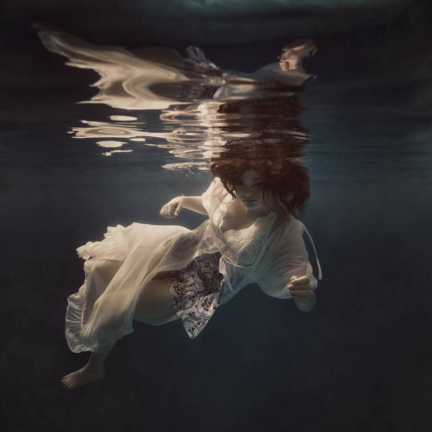 A girl in a light fluttering dress swims underwater as if floating in weightlessness against a dark background - Photo, image