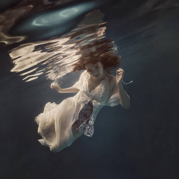 A girl in a light fluttering dress swims underwater as if floating in weightlessness against a dark background - Photo, image