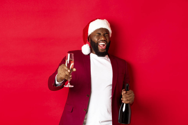 Χριστούγεννα, πάρτι και γιορτές. Αστείος μεθυσμένος μαύρος γιορτάζει την Πρωτοχρονιά, γελώντας και διασκεδάζοντας, κρατώντας ποτήρι και μπουκάλι σαμπάνιας, κόκκινο φόντο - Φωτογραφία, εικόνα