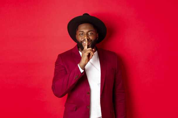 Winter holidays and shopping concept. Handsome Black man sharing secret, smiling and hushing at you with finger pressed to lips, standing against red background - Foto, immagini