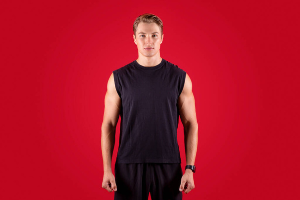 Selbstbewusster junger Mann in schwarzem T-Shirt und kurzer Hose blickt in die Kamera auf rotem Studiohintergrund - Foto, Bild