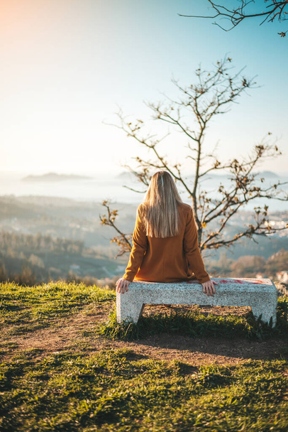 Vrouw op een gele jas zittend op een bankje met uitzicht op het landschap van Vigo, Galicië - Foto, afbeelding