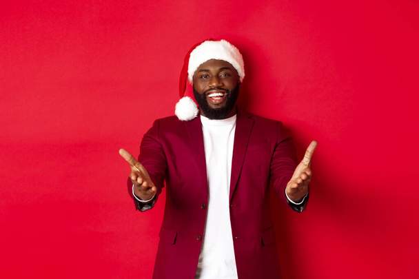Karácsony, parti és ünnepek koncepció. Boldog fekete férfi télapó kalap üdvözöl, mutató kezét a kamera elégedett mosollyal, álló ellen piros háttér - Fotó, kép