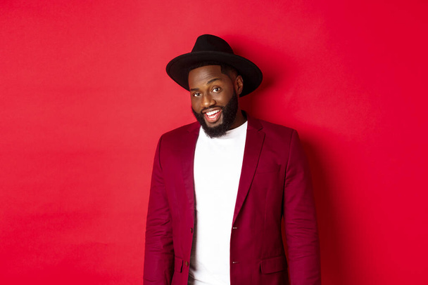 Christmas shopping and people concept. Handsome bearded african american guy looking at camera, smiling confident, wearing party clothes, red background - Photo, Image
