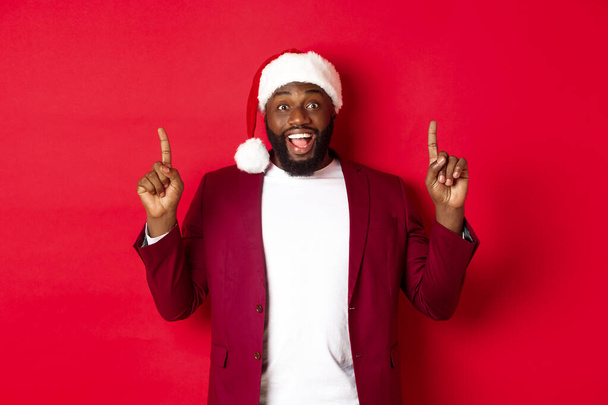 Karácsony, parti és ünnepek koncepció. Izgalmas fekete férfi télapó kalap és blézer, ujjal felfelé és mosolyogva, bemutató promóciós ajánlat, piros háttér - Fotó, kép