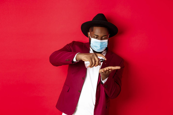 Covid-19, quarantine and holidays concept. Handsome Black man in face mask and party outfit, disinfecting hands with hand sanitizer, using antiseptic, standing over red background - Photo, Image
