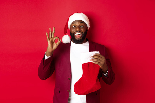 hombre negro asombrado celebración de regalos de vacaciones dentro de calcetín de Navidad, mostrando signo ok en la aprobación, como algo, de pie sobre el fondo rojo - Foto, imagen