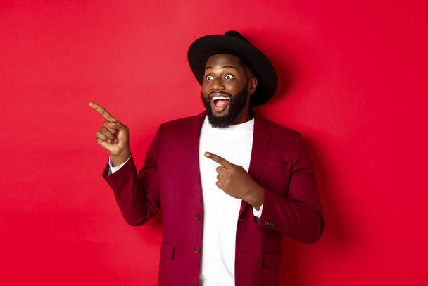 Winter holidays and shopping concept. Happy Black man pointing left and smiling, showing new year promo offer on red background - Foto, immagini