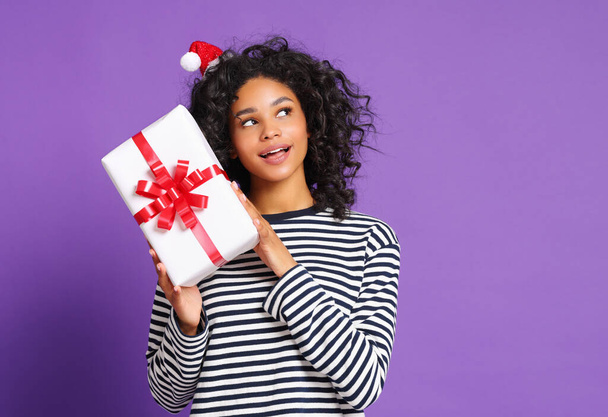 cheerful funny smiled young ethnic woman with santa hat looking away and shaking gift box near ear during Christmas celebration against colored purple backgroun - Foto, Imagen