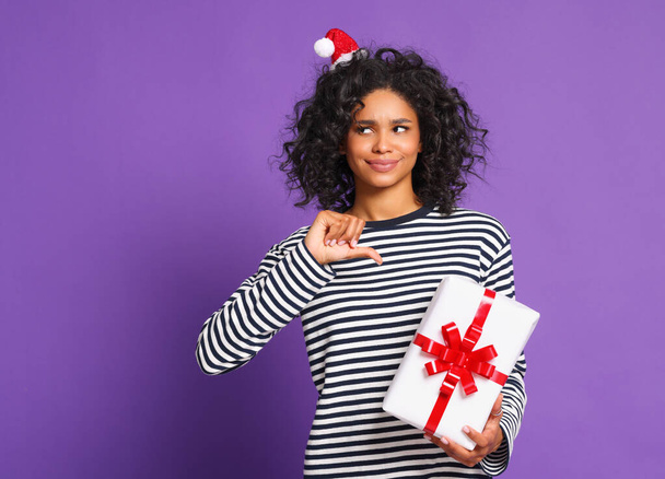 Dissatisfied young african american female in headband  outing lips and pointing at wrapped present during holiday celebration against purple backgroun - Foto, immagini