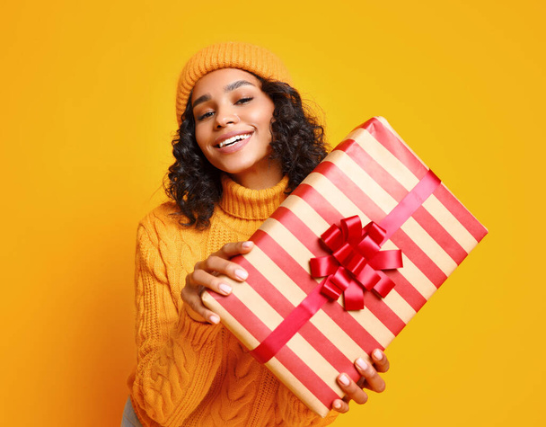 Szczęśliwa młoda etniczna kobieta w swetrze i kapeluszu pokazuje zapakowany prezent z uśmiechem na Boże Narodzenie na żółtym tle - Zdjęcie, obraz