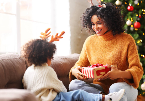 Feliz familia afroamericana étnica de vacaciones: madre alegre le da un regalo a su pequeño hijo lindo durante la celebración de Navidad en una acogedora habitación con tre - Foto, Imagen