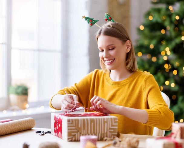 Очаровательная молодая женщина, улыбающаяся и обертывающая декоративную бумагу вокруг настоящей коробки, сидя за столом и готовясь к празднованию Рождества в доме - Фото, изображение