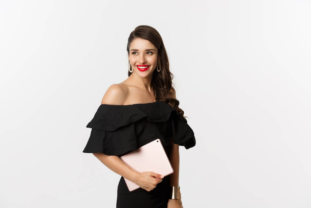 Fashion and shopping concept. Stylish young woman with glamour makeup, wearing black dress, holding digital tablet and smiling, white background - Фото, зображення