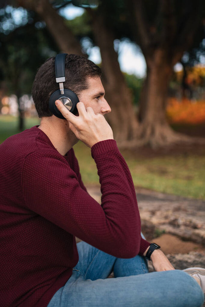 Blonde boy listening to music on wireless headphones sitting on the floor without looking at the camera. Enjoying the song. Musical concept. Red sweater. - Photo, Image