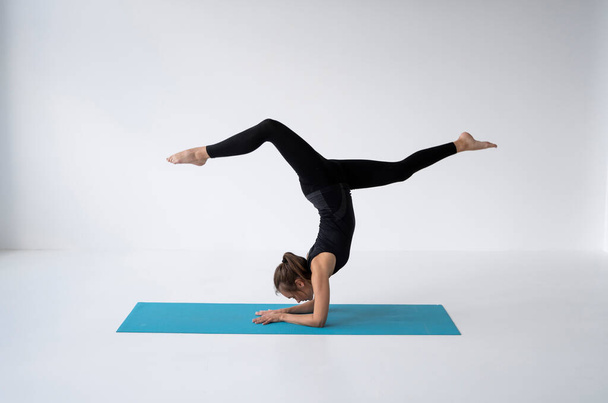 Young woman is practicing yoga in a studio. Slim young girl in a black sport wear is doing yoga practice on a blue mat. Concept of healthy life and natural balance between body and mental development. - Fotoğraf, Görsel