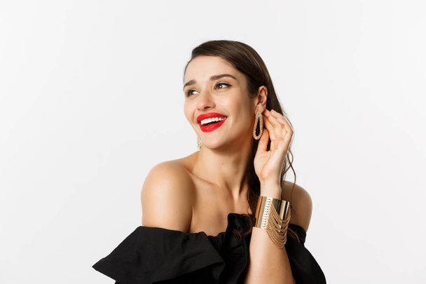 Fashion and beauty concept. Close-up of elegant brunette woman with red lips, black dress, laughing coquettish and gazing away, standing over white background - Photo, Image