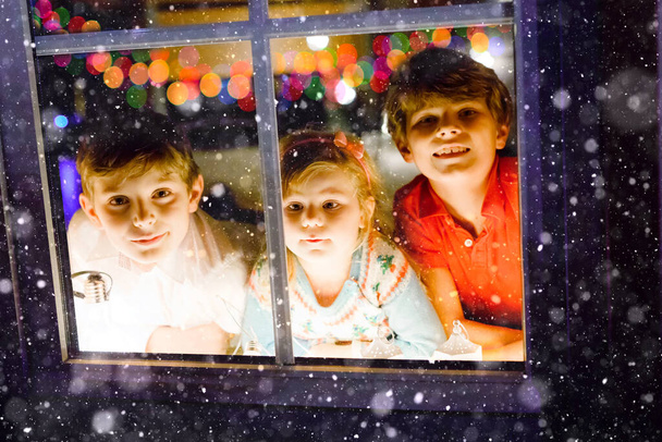 Trójka uroczych dzieci siedzących przy oknie w Wigilię. Dwóch chłopców ze szkoły i mała dziewczynka, rodzeństwo wyglądające na zewnątrz i marzące. Szczęście rodzinne na tradycyjnym urlopie - Zdjęcie, obraz
