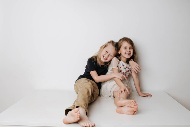 Happy siblings sitting together on table. Boy hugs girl, she leans away a bit. - Foto, imagen