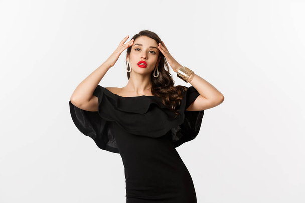 Confident and stylish woman in black elegant dress, looking sassy at camera, standing over white background - Photo, Image