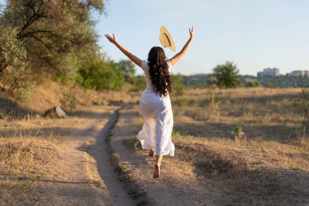 young girl in a white dress jumping in the field, tossing her hat up. Happy girl on a summer evening, against the backdrop of the setting sun Beautiful young woman wearing a white dress and hat Jumping jumping in the grass - Photo, Image