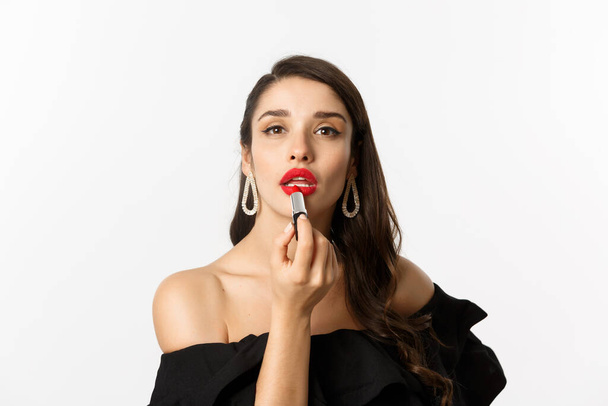 Close-up de mulher bonita aplicando batom vermelho nos lábios, olhando para a câmera como espelho, de pé em vestido preto sobre fundo branco - Foto, Imagem